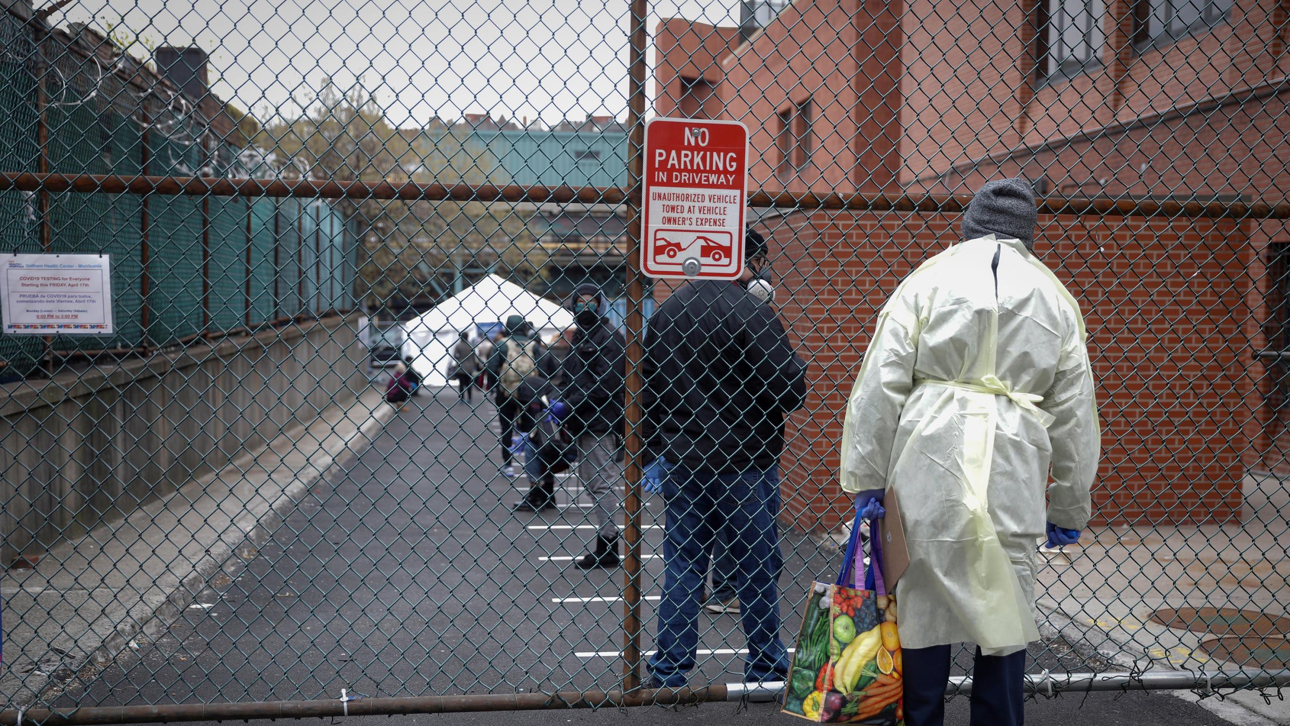 The photo shows a health worker standing behind a fence looking in on an enclosed parking lot where people are lined up awaiting testing.
